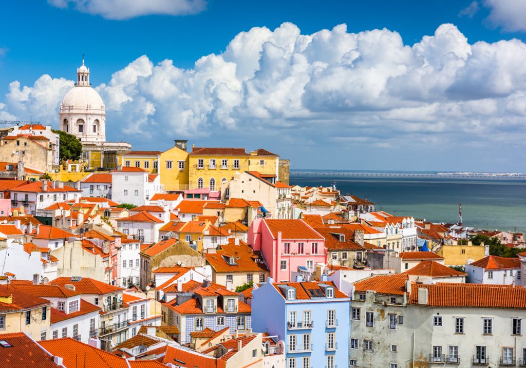 View of Lisbon in Portugal with mainly white buildings with red tiled roofs and a crowning white domed church towering above with the sea in the background