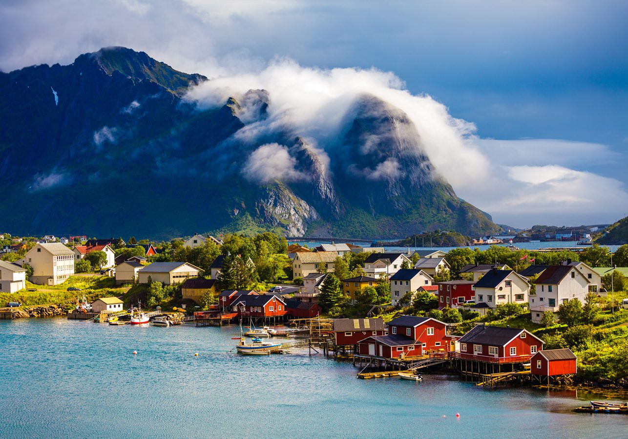 Red and white houses located in the coastal town of Lofoten in Norway with a cloud topped mountain in the background and calm see in the foreground